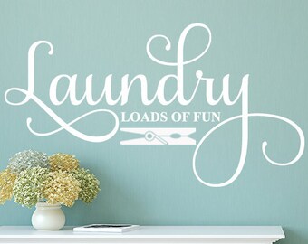 Puede incluir: Un adhesivo de pared blanco con las palabras "Laundry Loads of Fun" en una escritura decorativa. El adhesivo incluye un gráfico de pinza de ropa. Está en una pared turquesa sobre un mueble blanco con un jarrón de flores.