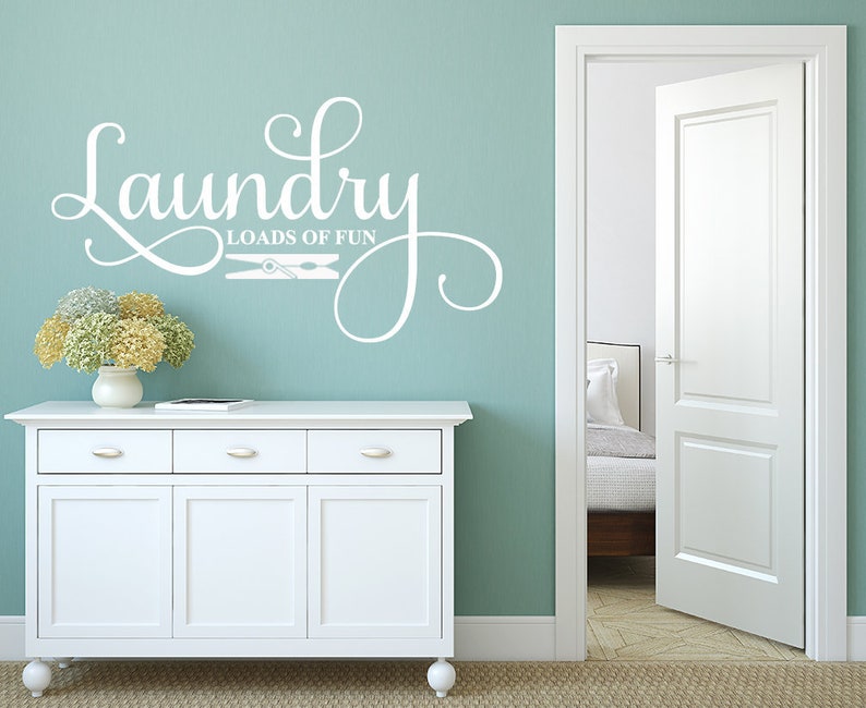 May include: A white wall decal with the words "Laundry Loads of Fun" in a decorative script. The decal includes a clothespin graphic. The decal is on a teal wall above a white cabinet with a vase of flowers.