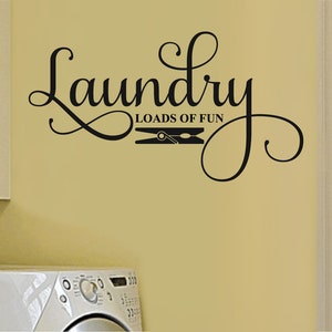 May include: Black wall decal with the words "Laundry Loads of Fun" in a decorative script font. A clothes pin graphic is between the words. The decal is on a light yellow wall, with a white washing machine and cabinet visible.