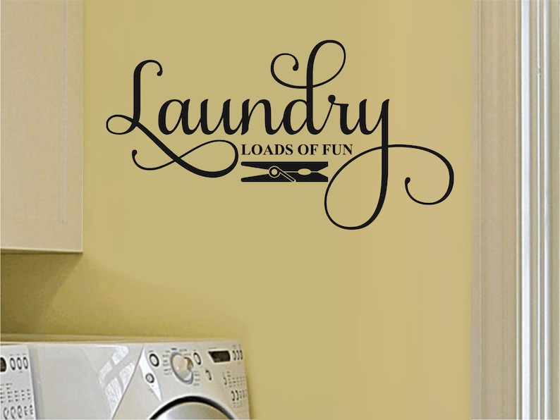 May include: Black wall decal with the words "Laundry Loads of Fun" in a decorative script font. A clothes pin graphic is between the words. The decal is on a light yellow wall, with a white washing machine and cabinet visible.