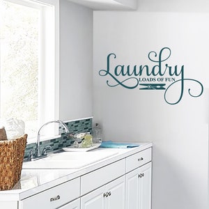 May include: A teal laundry room wall decal with the words "Laundry Loads of Fun" in a decorative script font. The design includes a clothespin graphic. The decal is affixed to a white wall in a laundry room setting.
