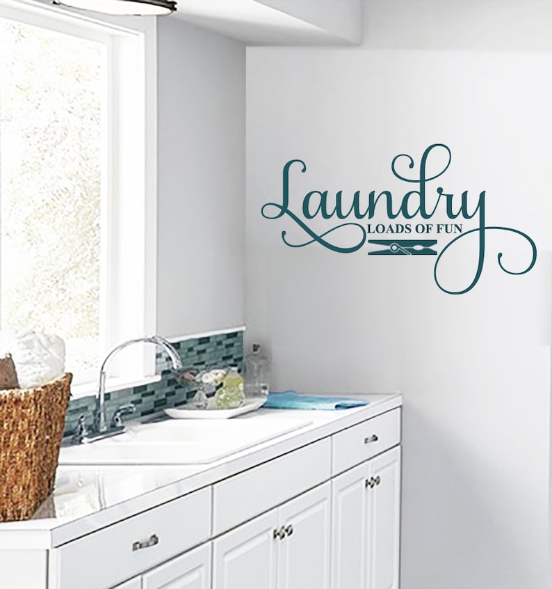 May include: A teal laundry room wall decal with the words "Laundry Loads of Fun" in a decorative script font. The design includes a clothespin graphic. The decal is affixed to a white wall in a laundry room setting.