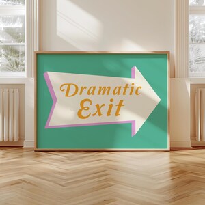 May include: A framed art print featuring a teal background with a white arrow pointing right. The arrow has a pink border and contains the words "Dramatic Exit" in a gold, stylized font. The frame is a natural wood color.