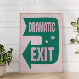 May include: A framed art print with a pink background and a green arrow-shaped design. The design features the words "DRAMATIC EXIT" in pink and green, with two pink stars. The print is in a wooden frame.