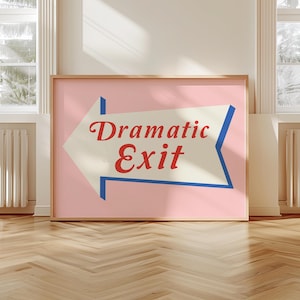 May include: A framed poster with a pink background and a white arrow pointing left. The arrow has a blue border and the words "Dramatic Exit" in red script. The poster is in a light wood frame and is displayed in a room with hardwood floors.