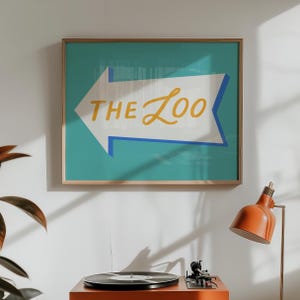 May include: A framed art print with a teal background and a white arrow pointing left. The text "THE ZOO" is written in yellow script within the arrow. The frame is a light brown color.