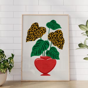 Puede incluir: Una impresión artística enmarcada que presenta una planta estilizada en un jarrón rojo en forma de corazón. La planta tiene hojas verdes grandes y hojas con estampado de leopardo. El fondo es blanco con un patrón de cuadrícula.
