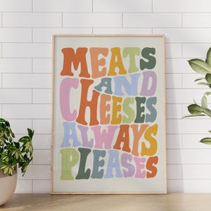 May include: A framed print with the words "MEATS AND CHEESES ALWAYS PLEASES" in a retro font. The text is in various colors, including orange, pink, green, and blue, against a cream background. The frame is a light wood color.