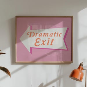 May include: A framed art print with a pink background and a white arrow pointing left. The arrow contains the words "Dramatic Exit" in orange script. The frame is a light wood color.