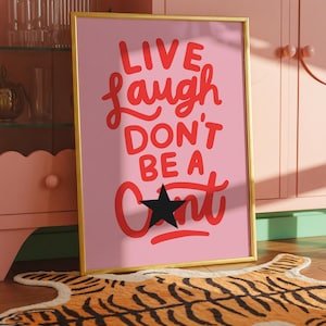 以下が含まれることがあります： 「LIVE, LAUGH, DON'T BE A」という赤い筆記体の言葉が書かれた、額入りのピンクのアートプリント。文字の「U」の代わりに黒い星が使われています。フレームは金色です。プリントは、トラ柄のラグが敷かれた木の床に飾られています。