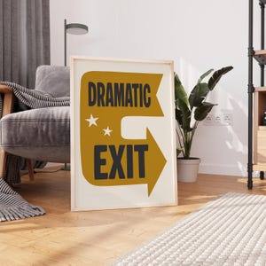 Puede incluir: Una impresión artística enmarcada con las palabras "DRAMATIC EXIT" en negro y dorado. El diseño incluye una flecha y dos estrellas. La impresión se muestra en un suelo de madera, apoyada contra una pared.