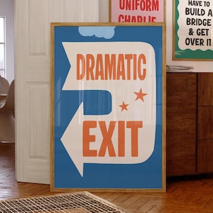 May include: A framed poster with a blue background and the words "DRAMATIC EXIT" in orange, with an arrow pointing left. Two orange stars and a cloud graphic are also present. The poster is in a wooden frame.