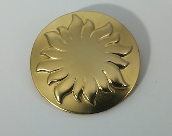 May include: A gold-colored metal brooch in the shape of a stylized sun with a raised design.