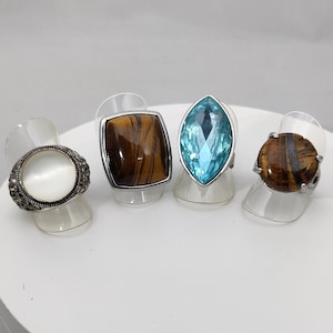 Puede incluir: Cuatro anillos con piedras de diferentes colores. Los anillos están expuestos en un soporte circular blanco. Las piedras son un óvalo blanco, un rectángulo marrón, una marquesa azul y un óvalo marrón.
