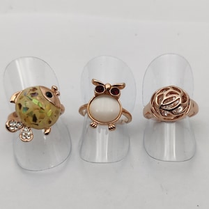 May include: Three gold-tone rings with different designs. The first ring features a green and white owl with red eyes. The second ring has a white oval stone with a gold-tone owl design. The third ring is a gold-tone circle with a delicate design.