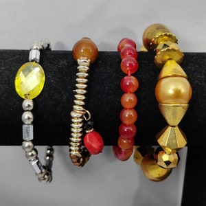 May include: Five bracelets stacked on a black display stand. The bracelets are made of different materials, including beads, metal, and stones. The bracelets are in shades of gold, silver, red, and yellow.