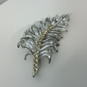 Peut inclure: Une broche en forme de plume argentée avec une rangée de strass clairs au centre.