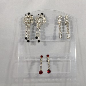 Peut inclure: Trois paires de boucles d'oreilles pendantes présentées sur un présentoir en plastique transparent. La paire du haut présente un motif en strass noir et blanc, la paire du milieu présente un motif en strass transparent et la paire du bas présente un motif en strass rouge.