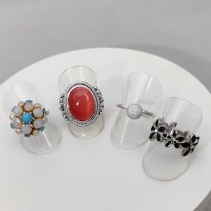 May include: Four silver rings on display stands. The rings feature different designs, including a red oval stone, a cluster of turquoise and white stones, a small round stone, and a floral design with black stones.