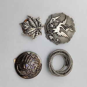 Puede incluir: Cuatro broches de metal plateados con diferentes diseños. Uno presenta una hoja de arce, otro un pájaro en vuelo, un tercero un animal estilizado y el último es un círculo trenzado.