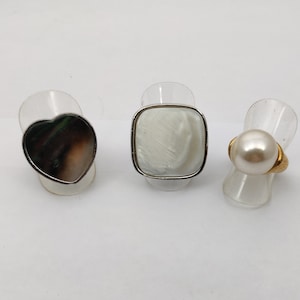 May include: Three rings on display stands. The ring on the left has a heart-shaped black and brown stone. The middle ring has a square white stone. The ring on the right has a white pearl.