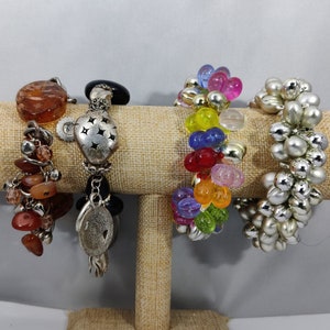 May include: Five bracelets displayed on a brown fabric stand. The bracelets are made of various materials, including beads, metal, and stones. The bracelets are in different colors, including brown, silver, and rainbow.