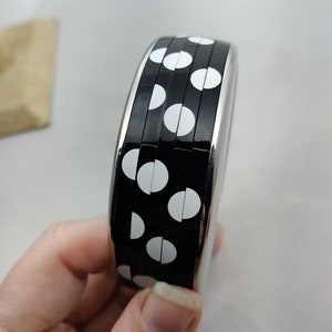 Könnte beinhalten: Ein schwarz-weiß gepunktetes Armreif-Armband. Das Armband hat eine glänzende schwarze Oberfläche mit weißen Punkten und eine silberfarbene Metallverkleidung. Das Armband wird in einer Hand gehalten.