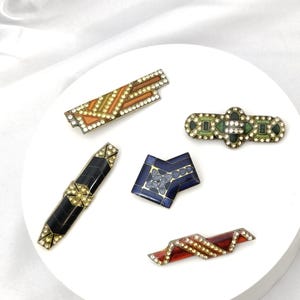 Op de afbeelding: Een verzameling vintage broches in diverse geometrische vormen. De broches hebben een mix van kleuren, waaronder zwart, rood, oranje en blauw, geaccentueerd met heldere strass-steentjes. De broches worden gepresenteerd op een wit oppervlak.
