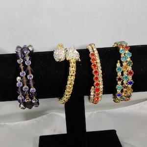 Peut inclure: Quatre bracelets décoratifs exposés sur un présentoir noir. Les bracelets présentent divers motifs, notamment des perles violettes, de l'or avec des pierres transparentes, des pierres rouges et des pierres colorées. Les bracelets sont posés sur un fond blanc.