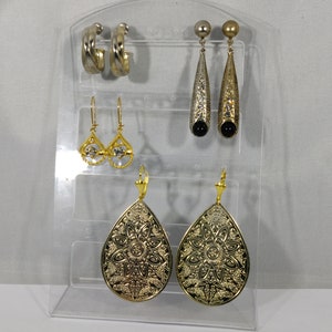 Könnte beinhalten: Ein Set mit sechs Ohrringen, die auf einem durchsichtigen Acrylständer ausgestellt sind. Die Ohrringe sind in Gold- und Silbertönen gehalten. Zwei Paar Ohrringe haben eine Tropfenform mit filigranen Designs. Ein Paar Ohrringe ist im einfachen Creolen-Stil gehalten. Das restliche Paar Ohrringe hat eine Tropfenform mit einem schwarzen Stein am unteren Ende.