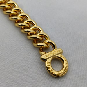 Peut inclure: Un bracelet chaîne doré avec de larges maillons arrondis. Le fermoir porte l'inscription "PARFUMS GUY LAROCHE PARIS" en cercle. Le bracelet présente une finition brillante et polie.