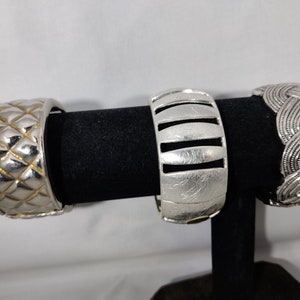 May include: Three silver metal bracelets with different designs. The bracelet on the left has a quilted pattern, the middle bracelet has a vertical striped pattern, and the bracelet on the right has a braided pattern.
