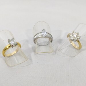 May include: Three rings with clear gemstones on display stands. The rings are gold and silver with different styles of settings.