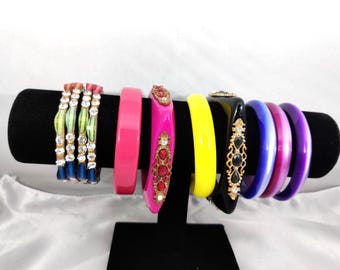 Puede incluir: Una colección de nueve pulseras tipo brazalete en un soporte negro. El conjunto incluye tres brazaletes a rayas azules y verdes con piedras transparentes, un brazalete rosa sólido, uno rosa con piedras rojas, uno amarillo y tres morados.