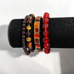 May include: Four beaded bracelets in shades of red, brown, and gold are displayed on a black velvet surface. The bracelets feature various bead shapes and sizes, including round, faceted, and rectangular beads. The bracelets are a mix of materials.