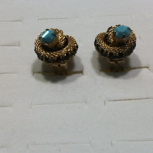 May include: A pair of vintage clip-on earrings. Each earring features a gold-toned, woven design with a central turquoise-colored stone and a border of small, dark-colored stones. The earrings are displayed on a white surface.