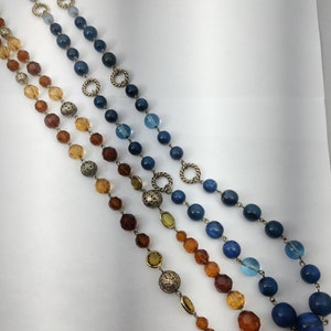 Può includere: Tre fili di collane di perle con perle blu, blu chiaro e ambra. Le collane sono sovrapposte e presentano accenti metallici decorativi.