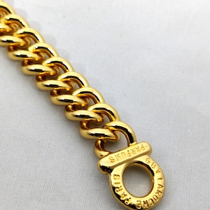 Op de afbeelding: Een goudkleurige metalen kettingarmband met dikke, in elkaar grijpende schakels. Het uiteinde van de ketting eindigt in een ronde sluiting gegraveerd met de tekst GUY LAROCHE PARIS PARFUMS, met een gepolijste en reflecterende afwerking.