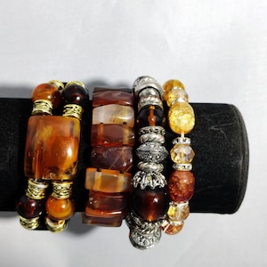 May include: Four beaded bracelets in shades of brown, amber, and gold. The bracelets feature a variety of beads, including large rectangular and round beads, and silver accents. The bracelets are displayed on a black surface.