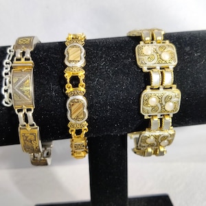 May include: Three decorative bracelets in gold and silver tones, displayed on a black stand. The bracelets feature detailed patterns and decorative elements. One has a chain, the others have rectangular and oval links.