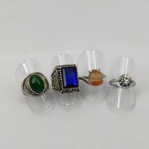 Peut inclure: Quatre bagues en argent avec des pierres précieuses de différentes couleurs. Les bagues sont présentées sur des supports à bagues en plastique transparent. Les bagues présentent une pierre ovale verte, une pierre rectangulaire bleue, une pierre ovale orange et une pierre ronde transparente.