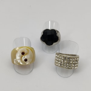 Peut inclure: Trois bagues en argent avec des motifs différents. La première bague a une pierre noire en forme de fleur. La deuxième bague a une coquille blanche avec un symbole yin-yang et deux points rouges. La troisième bague a une bande en argent avec des strass transparents.