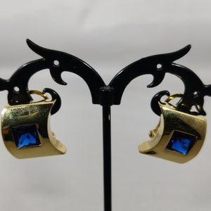 Puede incluir: Un par de pendientes de clip de tono dorado con piedras cuadradas azules engastadas en el centro de cada pendiente.