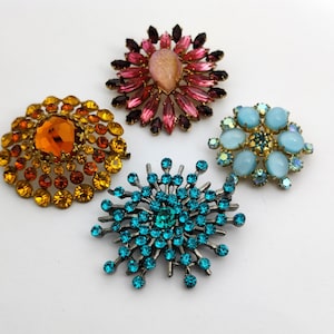 Op de afbeelding: Vier vintage broches met kleurrijke strass accenten. De broches hebben verschillende vormen, waaronder een bloem, een steruitbarsting en een cirkelvormig ontwerp. Kleuren zijn onder meer oranje, geel, roze, blauw en blauwgroen. Deze decoratieve broches zijn perfect.