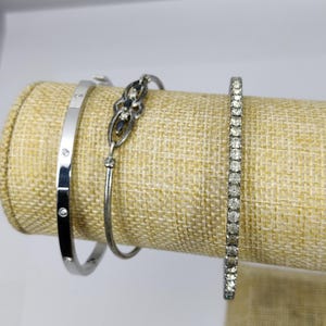 Puede incluir: Tres pulseras de plata con diseños diferentes. La pulsera de la izquierda tiene una fila de pequeños diamantes. La pulsera del medio tiene un diseño delicado y adornado con piedras negras. La pulsera de la derecha tiene una fila de diamantes más grandes.