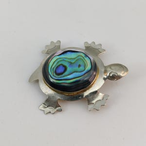 Peut inclure: Broche argentée en forme de tortue avec un centre en coquillage d'ormeau aux couleurs vives. Le coquillage présente des reflets bleus, verts et violets. La broche a un design détaillé avec des pattes, une tête et une queue visibles.