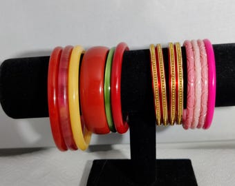 Può includere: Una collezione di braccialetti colorati esposti su un supporto nero. I braccialetti sono in varie tonalità di rosso, giallo, verde e rosa. Alcuni hanno dettagli dorati e motivi intagliati, mostrando una varietà di stili.