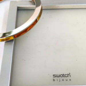 Könnte beinhalten: Ein silberner Bilderrahmen mit weißem Innenraum und dem Text "swatch bijoux" in Schwarz. Ein goldfarbenes Armband mit orangefarbenen und gelben Akzenten ist teilweise in der oberen linken Ecke sichtbar.
