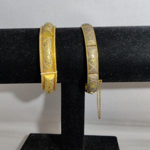 Peut inclure: Deux bracelets dorés à charnière avec des motifs gravés complexes. L'un présente un motif floral détaillé, l'autre un motif géométrique. Les deux sont exposés sur un présentoir en velours noir, mettant en valeur leur artisanat orné.