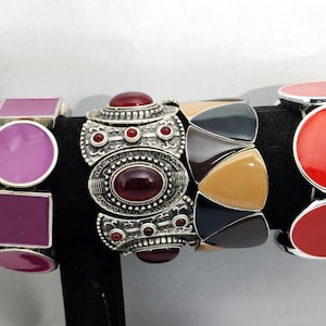 May include: Three stretch bracelets with various designs. One bracelet features a silver setting with a large oval red stone and smaller red stones. Another has geometric shapes in black, tan, and gray. The third has pink and purple shapes.